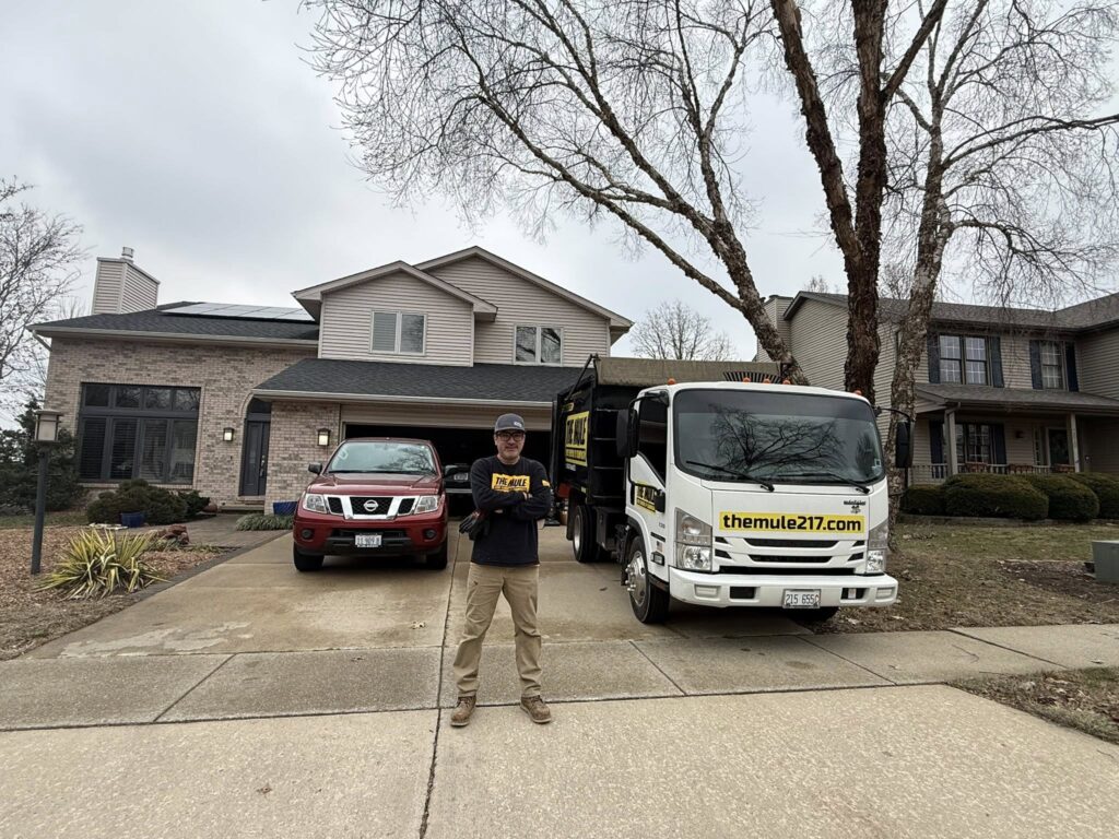 Man standing in driveway in front of junk removal truck and pickup truck outside suburban home