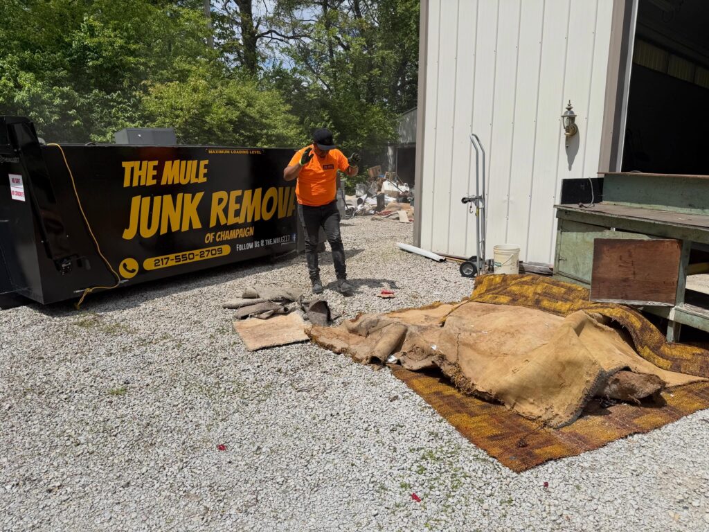 A worker in an orange shirt standing next to piles of old rolled-up carpet and a large black dumpster branded with The Mule Junk Removal.