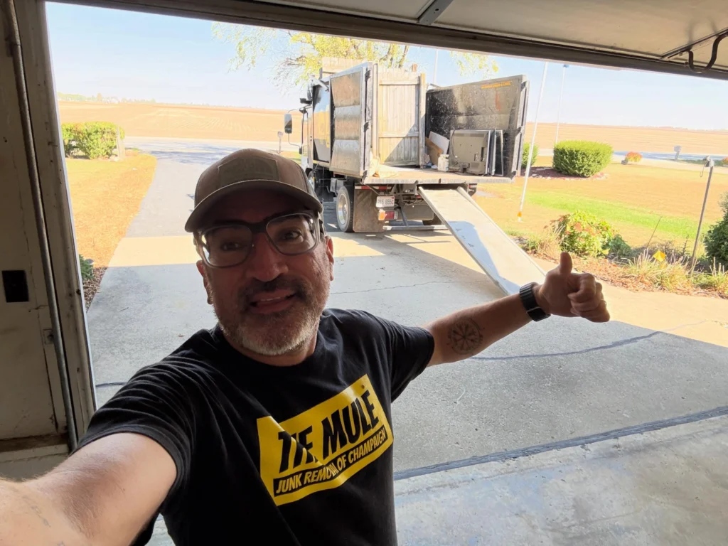 A smiling team member from The Mule Junk Removal of Champaign gives a thumbs-up in front of a junk removal truck with a ramp extended on a driveway during a property cleanout.