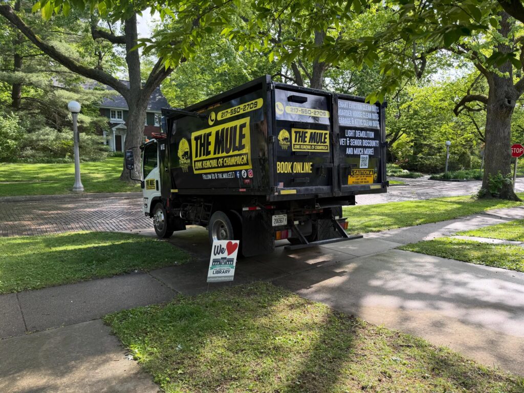 A branded black and yellow "The Mule Junk Removal of Champaign" truck parked on a residential brick street in Urbana, IL.