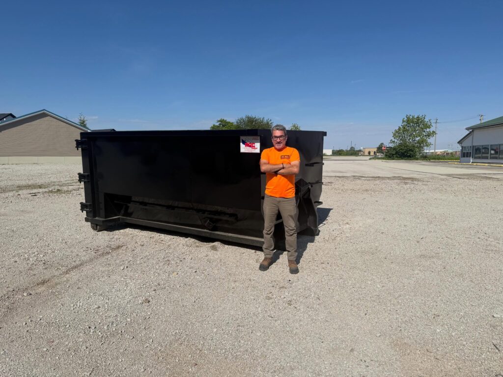 Team member standing with arms crossed next to a large, clean black roll-off dumpster in a gravel lot.