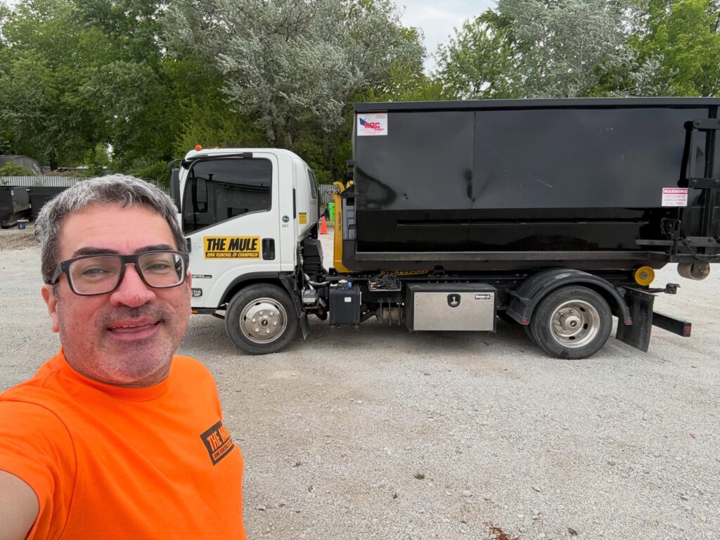 A man in an orange shirt and work gloves standing inside a dumpster with several stacked mattresses and old furniture.