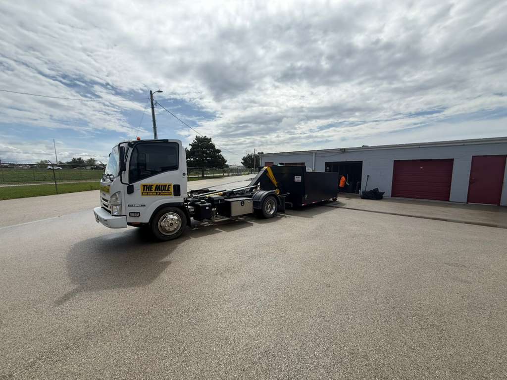 A commercial flatbed junk removal truck from The Mule parked in front of a row of outdoor storage units and garages during a cleanout service under a cloudy sky.