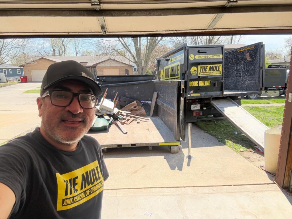 A team member from The Mule Junk Removal standing in a driveway next to a large black roll-off dumpster and a service truck with a loading ramp.