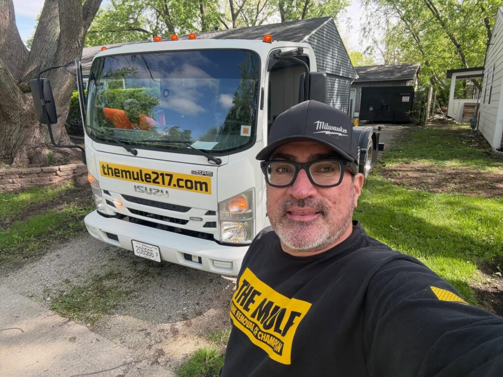 Owner of The Mule Junk Removal taking a selfie in front of a white branded Isuzu junk removal truck parked on a residential driveway in Champaign, IL.